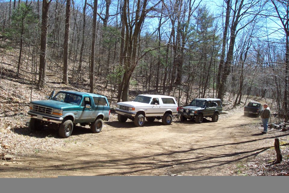 Teal Ford Bronco leads a line of white Bronco and Land Rover Discovery on a wooded dirt trail