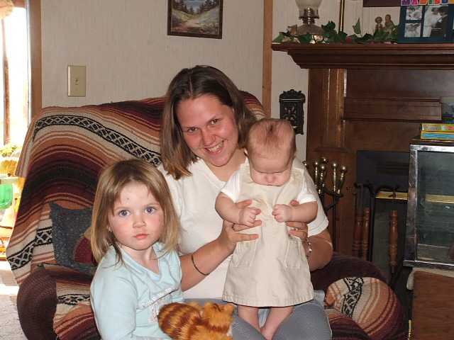 Woman smiles holding a baby while a young girl sits beside her on a couch