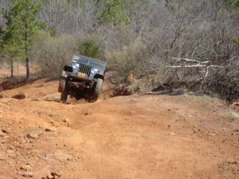 1995 Ford Bronco - Olive-drab flat-fender Willys-style Jeep dropping down a red-clay embankment at an off-road park, shot head-on. It runs a flatbed rear, exp…