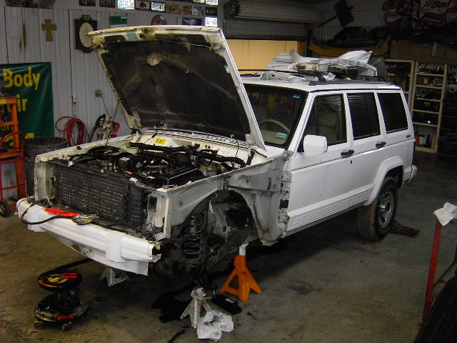 White Jeep Cherokee with hood open, front end disassembled, on jack stands in a garage