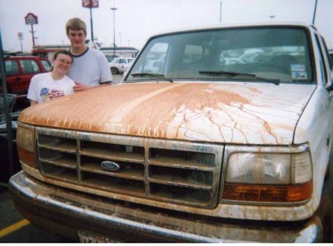 1995 Ford Bronco - Two people stand beside a white 1990s Ford Bronco in a parking lot, its hood, grille and bumper caked in dried orange-red mud after a wheel…