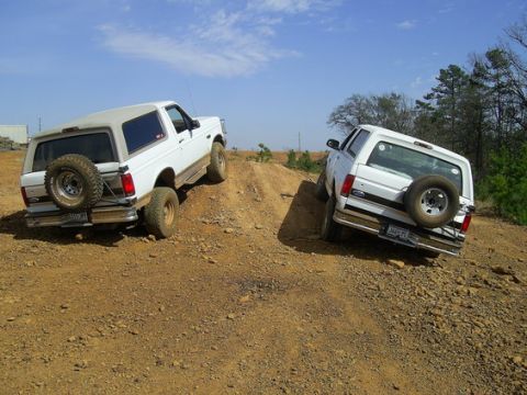 1995 Ford Bronco - Two white full-size Ford Bronco SUVs climbing opposite sides of a red-clay dirt mound at an off-road park, photographed from behind. Both c…