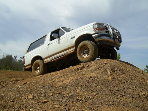 1995 Ford Bronco - Low-angle 3/4 front view of a white two-tone Ford Bronco full-size SUV climbing a red-clay dirt mound, shot from below against an overcast…