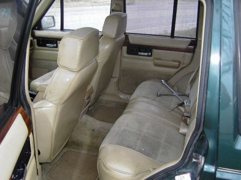 1993 Jeep Cherokee - The interior of a Jeep Cherokee shows tan seats and door panels, with wood trim accents and a floor jack secured by the seatbelt.