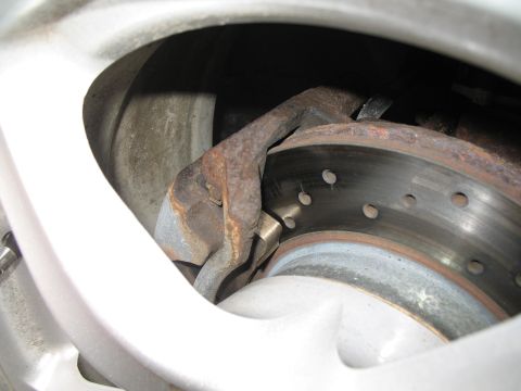 1996 Ford Probe - Close-up through the spokes of a silver alloy wheel at a cross-drilled brake rotor and its surface-rusted single-piston sliding caliper and…