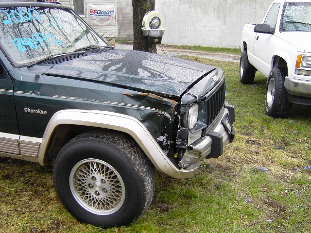 Front corner of a green 1993 Jeep Cherokee with damage to the fender and hood, parked next to a white pickup truck