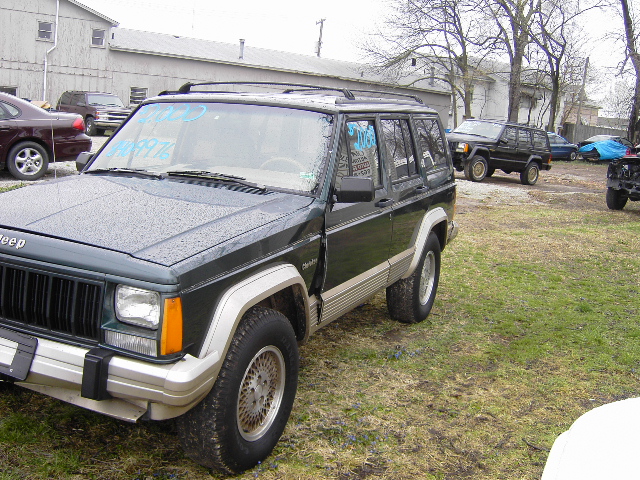 Dark green Jeep Cherokee parked outside with other vehicles in the background. Some writing on the windshield