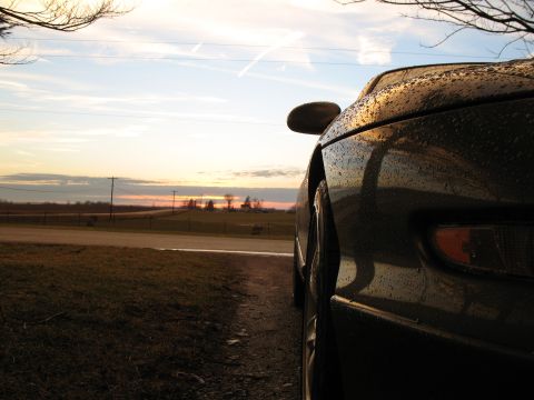 1996 Ford Probe - A dark car is seen from the side, covered in water droplets, as the sun sets on the horizon.