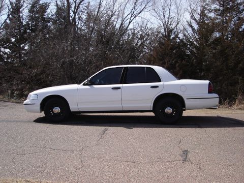 2000 Ford Crown Victoria - Side profile of a white 2000 Ford Crown Victoria sedan on a paved road with trees in the background.