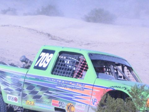Green off-road race truck number 709 with window net racing through the desert in a dust cloud