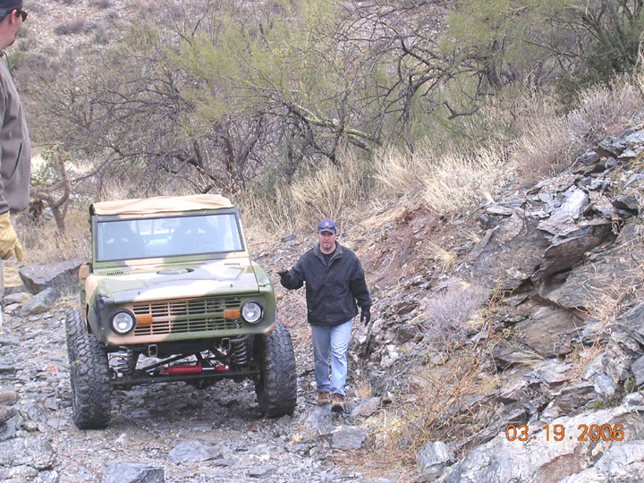 Green early Ford Bronco with tan soft top and red suspension links on a rocky desert trail