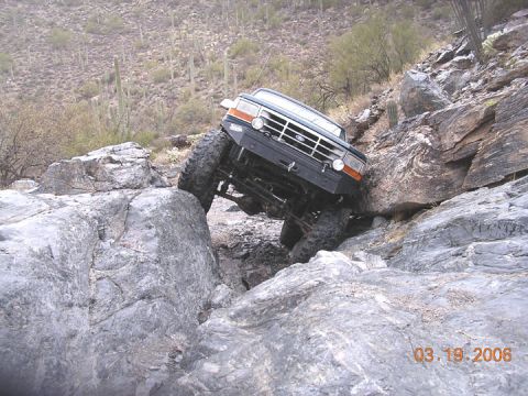 1994 Ford Bronco - A black 1994 Ford Bronco is shown off-roading through a rocky landscape, with its tires climbing over large boulders.