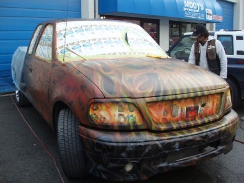 1985 Ford Bronco II - A pickup truck with a flame paint job is parked outside a garage, with its windows taped off for painting.