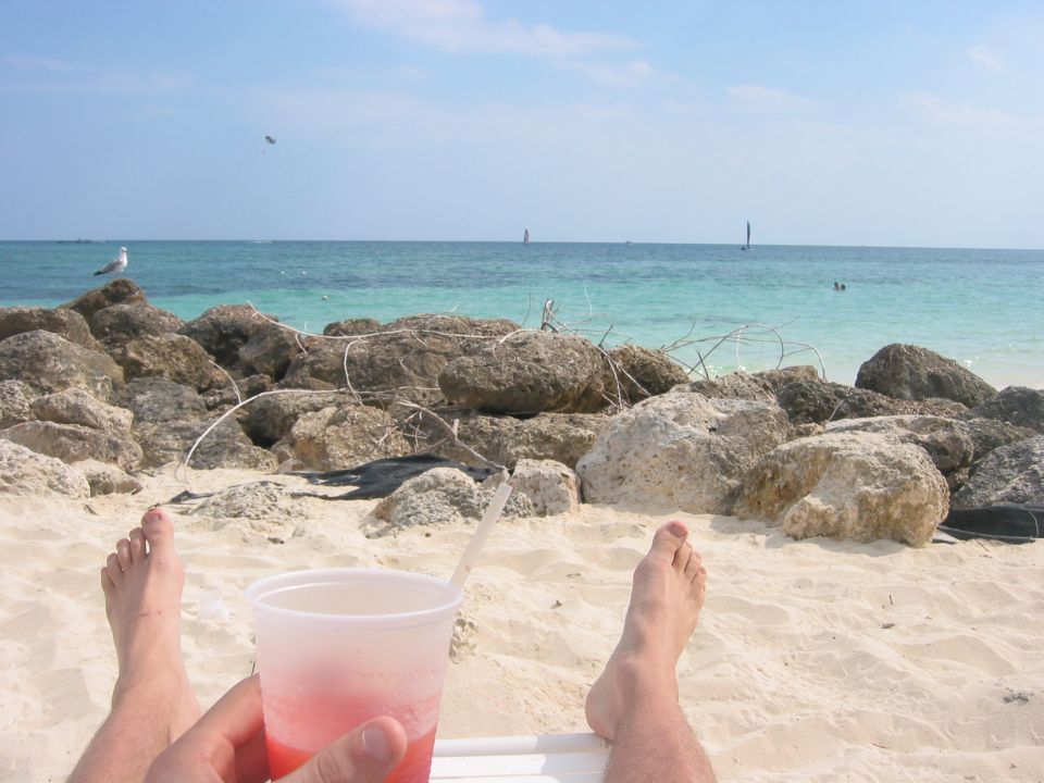 Beachgoer's bare feet and a pink drink with turquoise sea, rocks and gulls in view