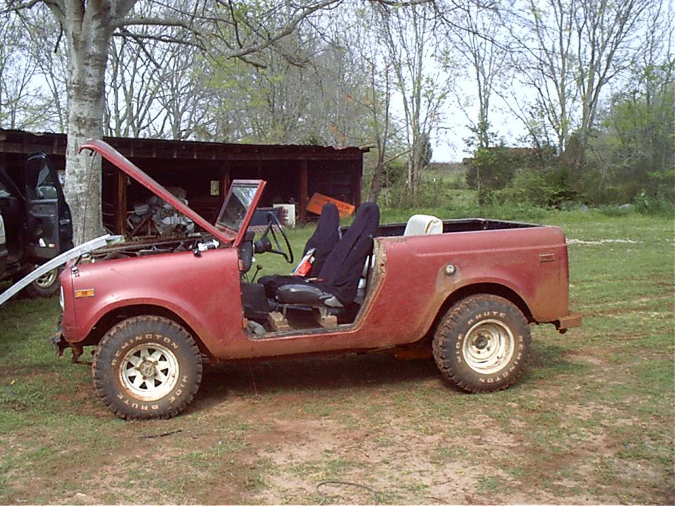 Faded red doorless vintage SUV with hood raised, white wheels and Remington tires, parked on grass by a shed