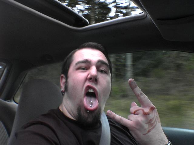 Man with tongue piercing and rock-on hand gesture inside a car with a sunroof