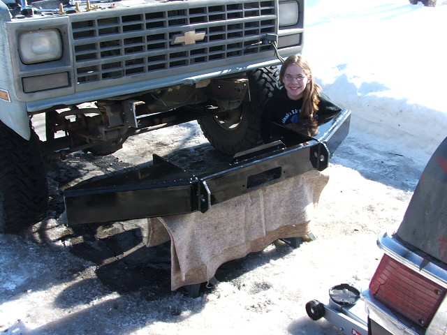 Squarebody Chevy front with new black steel bumper being fitted, person kneeling behind.