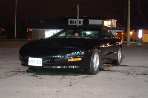 1981 Ford Mustang - A black coupe is parked outside a building at night, with its headlights on.