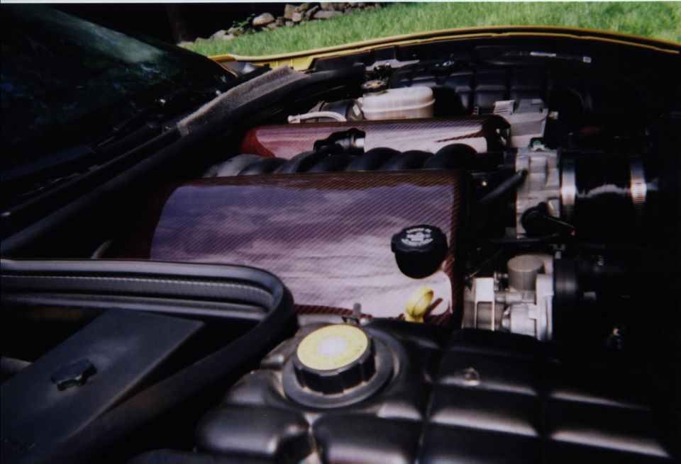 Engine bay of a yellow C5 Corvette showing LS1 V8 with red intake cover, hood open