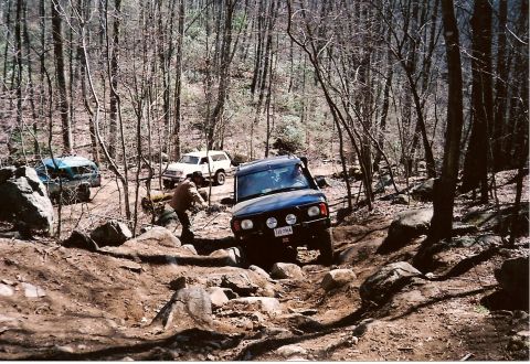 1987 Ford Bronco - Multiple SUVs traverse a challenging off-road trail through a wooded area.