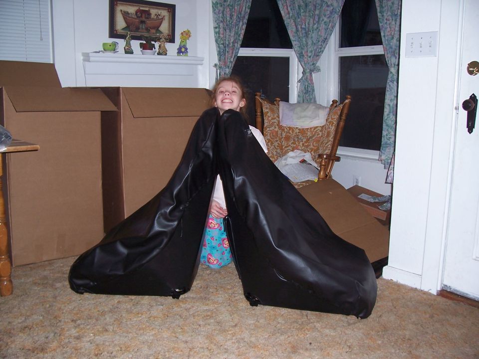 Two black car seats are positioned in front of large cardboard boxes, with a young girl sitting between them