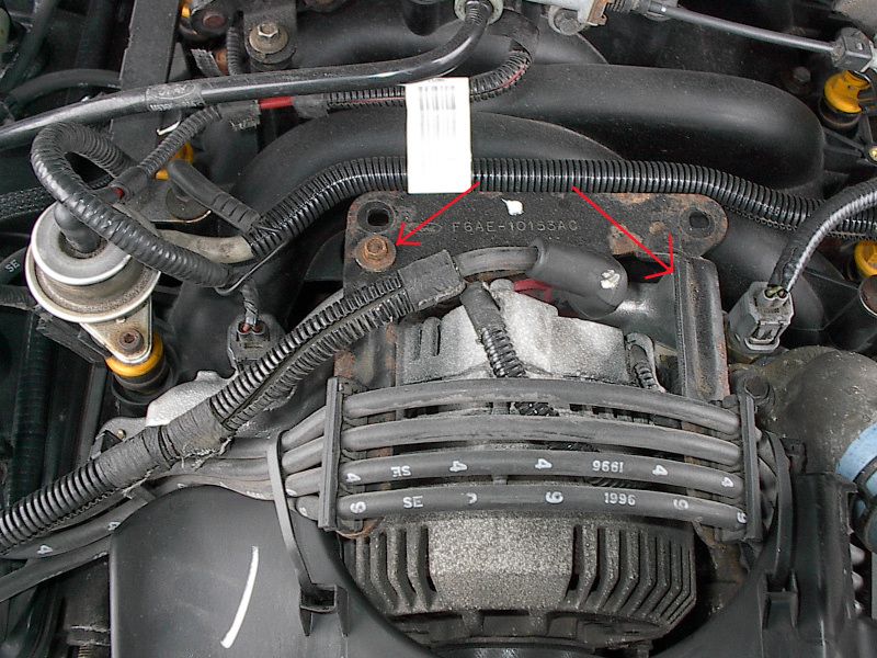 Engine intake manifold close-up with spark plug wires and red arrows pointing to bolts