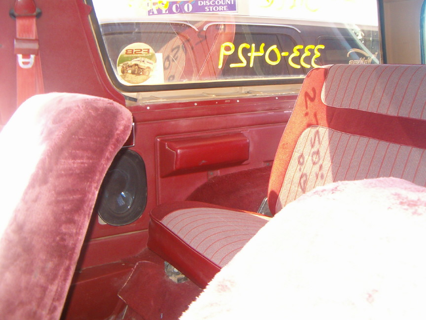 Interior view of a 1985 Ford Bronco showing red velour seats and door panel, with a sticker on the rear window