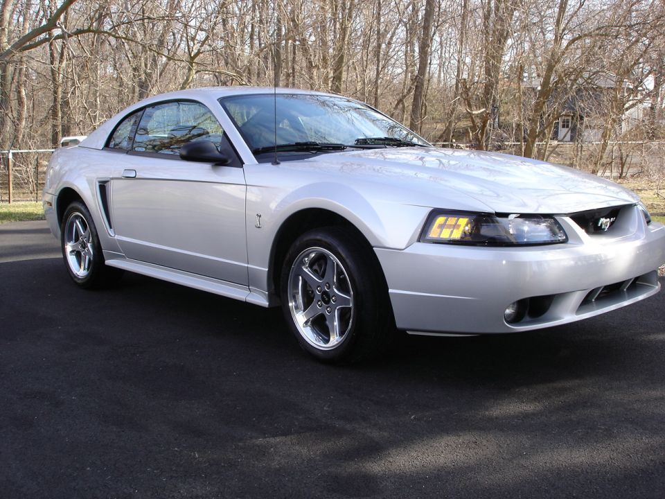 Silver Ford Mustang coupe parked on asphalt, trees in background. Front 3/4 view