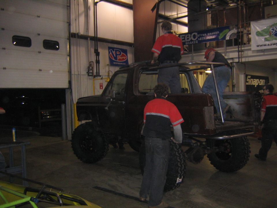 Dark SUV with custom roll cage and large tires, being worked on by people in red shirts