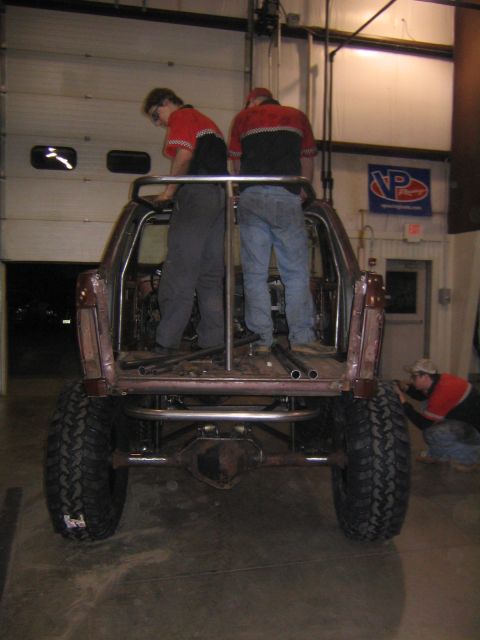 1978 Ford Bronco - Custom off-road vehicle build in progress with people working on the roll cage and rear tire.