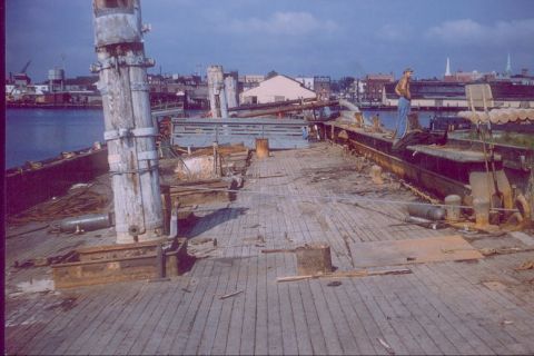1988 Ford Bronco - A man stands on the weathered wooden deck of a ship, surrounded by industrial equipment and a harbor in the background.