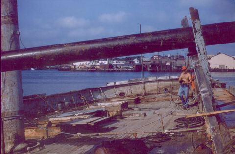 1988 Ford Bronco - A worker stands on the deck of a ship, with a large pipe overhead and a town visible across the water.