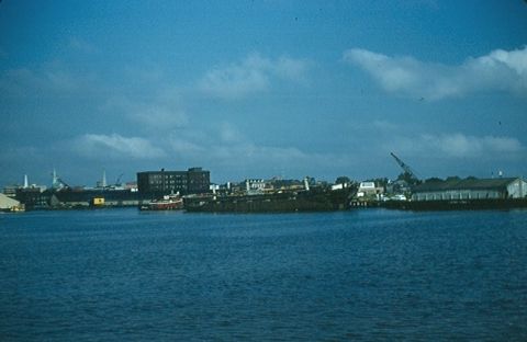 1988 Ford Bronco - A waterfront scene shows industrial buildings and ships docked along the water under a partly cloudy sky.