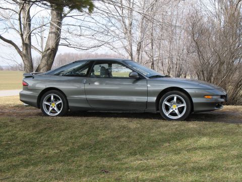 1996 Ford Probe - Side profile of a gray Ford Probe GT hatchback parked on grass in front of bare winter trees, with pop-up headlights closed, GT rocker badg…