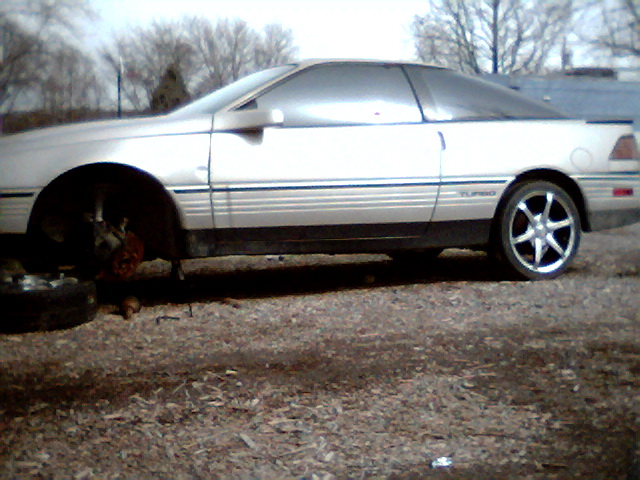 Silver coupe in profile, front wheel removed, jacked up on dirt lot