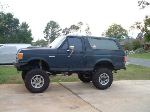 1995 Ford Bronco - This lifted Ford Bronco, likely from the early to mid-1990s, sits on a paved driveway with a suburban background.