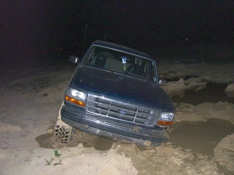 1992 Ford Bronco - A dark blue Ford Bronco is stuck in mud at night, with its front right tire sunk deep.