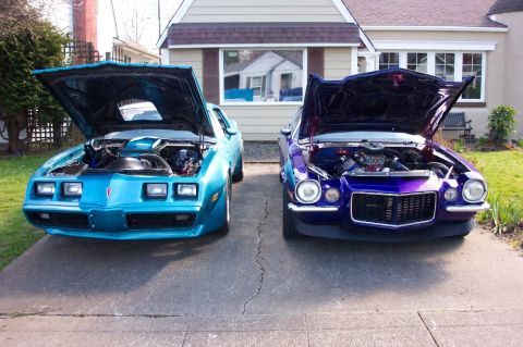 1979 Pontiac Trans Am - Two 1979 Pontiac Trans Ams, a teal one and a purple one, are parked in front of a house with their hoods open.