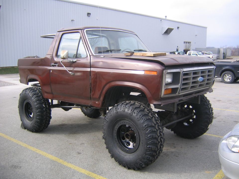 Brown topless lifted 1980s Ford Bronco on mud tires parked in front of a white metal building