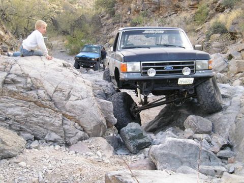1990 Ford Bronco - A dark blue Ford Bronco is shown off-roading on a rocky trail with another vehicle behind it.