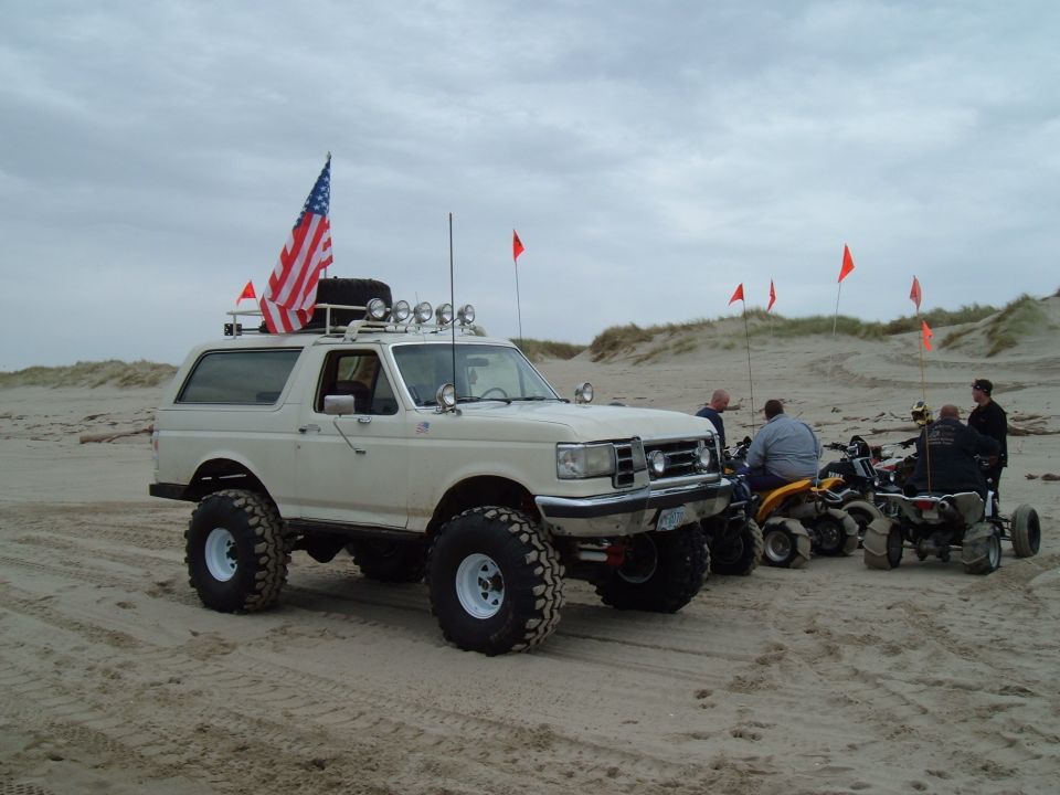 White lifted Ford Bronco with flag and light bar on beach beside ATVs