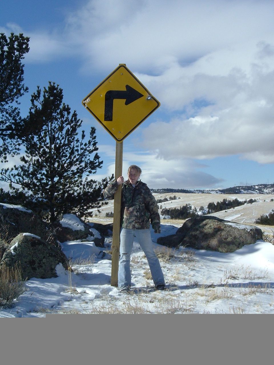 Person in camo jacket and jeans stands by a diamond-shaped yellow sign with a black arrow indicating a sharp right turn. Snowy, hilly landscape under a cloudy sky