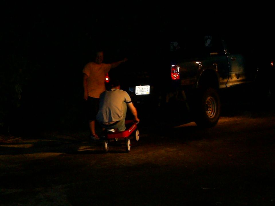 Person in red wagon pulled by another person next to a dark-colored truck at night