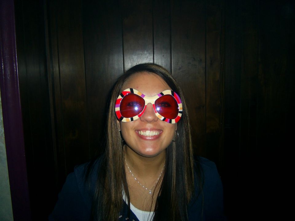 Woman with long brown hair and a wide smile wears large, round, colorful novelty sunglasses with red lenses
