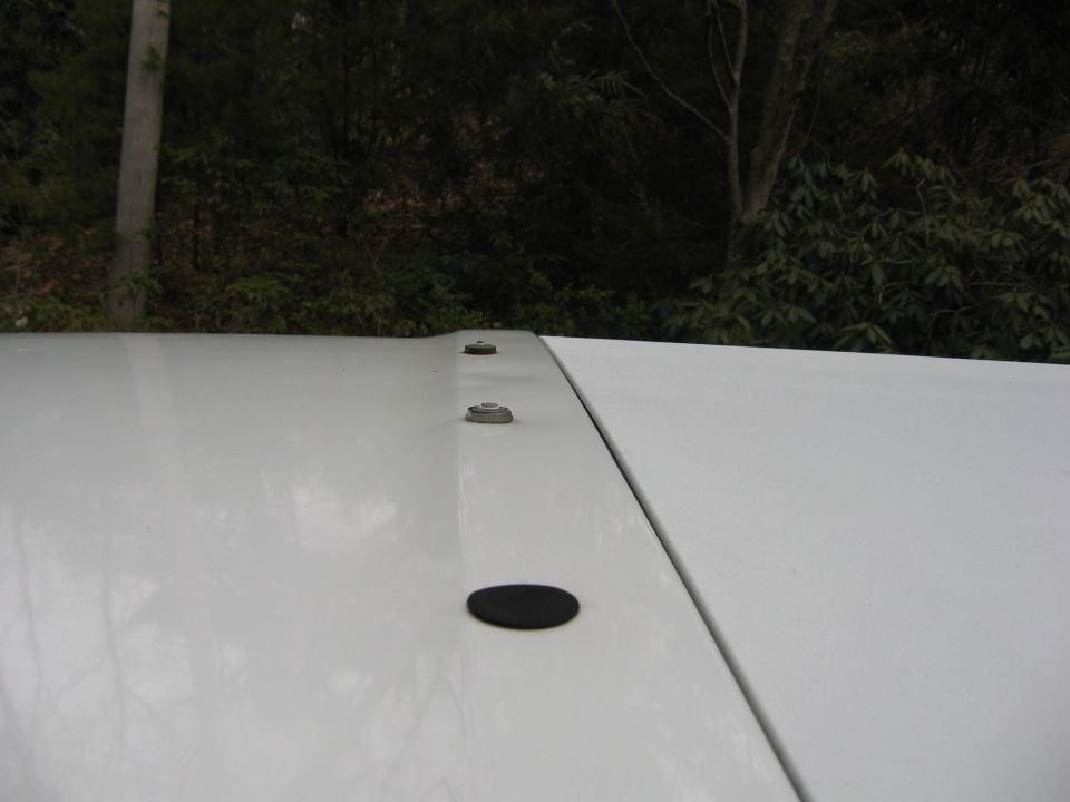 Close-up of white vehicle roof seam with bolts and rubber plug, trees in background
