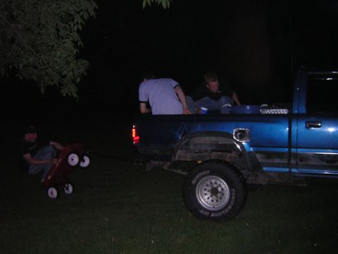 1985 Ford Bronco - People ride in the bed of a blue pickup truck while a red wagon is pulled behind it in the dark.