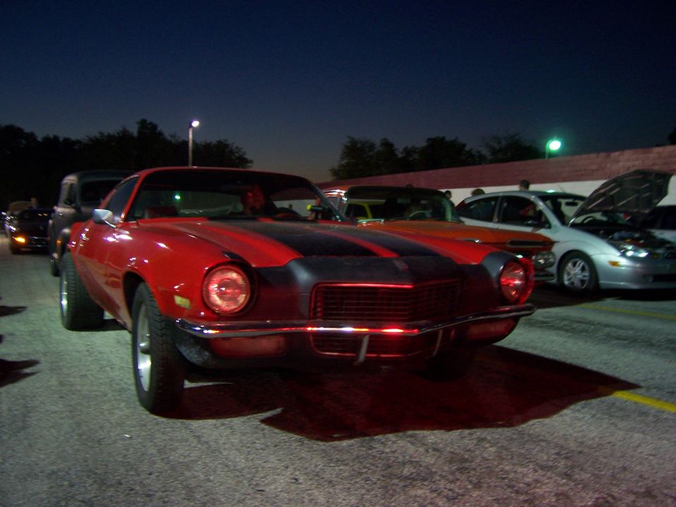 Front view of a red classic muscle car with black stripes. Other cars are parked behind it