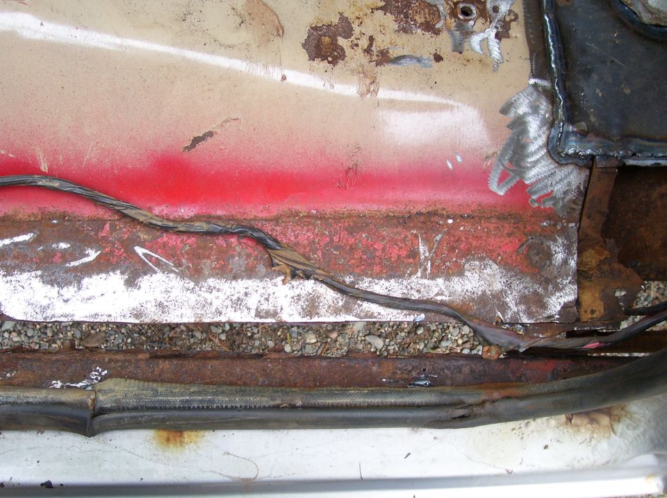 Close-up of a rusted vehicle floor pan with exposed wiring and gravel