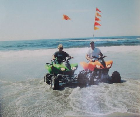 1994 Ford F-150 - Two people ride ATVs through the surf on a sunny day, with flags flying from poles attached to the vehicles.