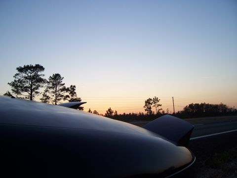 1994 Ford Probe - The front of a car is visible with pop-up headlights at dusk, with trees and a road in the distance.
